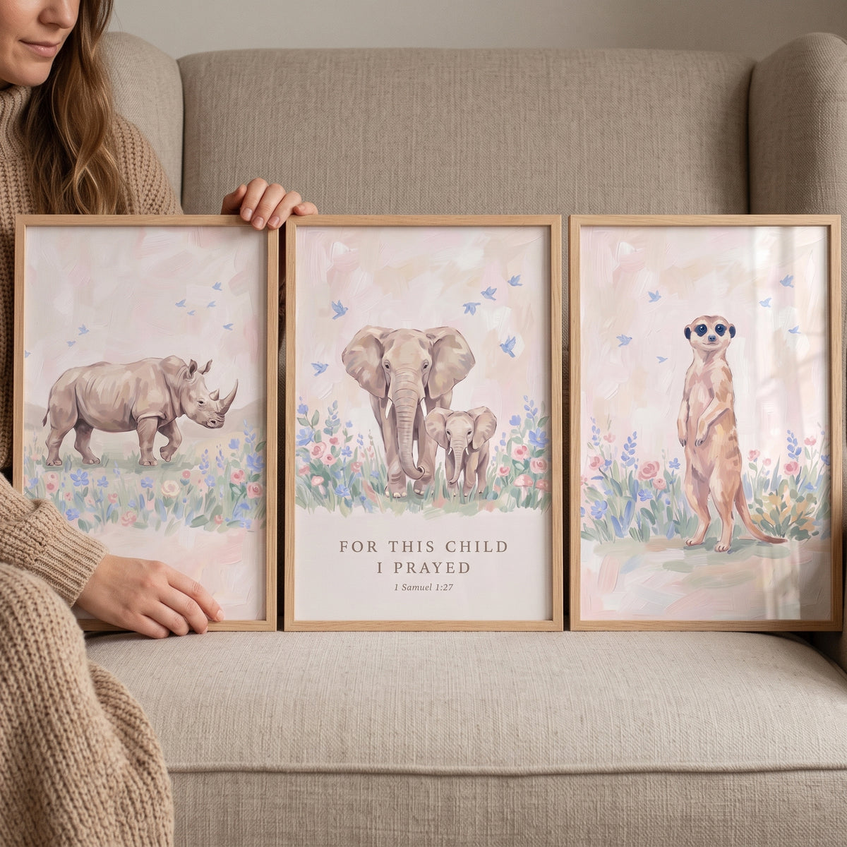 A person holds a pink meadow savanna animals wall art set of 3 featuring a rhino, elephants, and a meerkat on a couch.