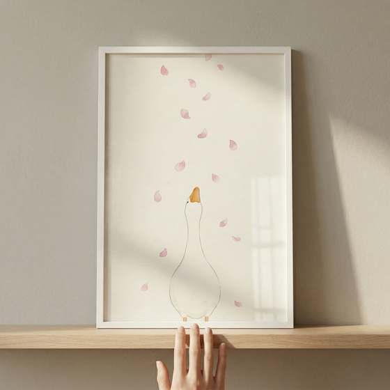 Whimsical minimalist art in a white frame sits on a wooden shelf. The illustration features a simple white goose from the back, looking up towards a shower of soft pink petals against a cream background. A person's hand is visible at the bottom, gently touching the frame's edge. Soft side lighting casts gentle shadows across the beige wall and the shelf.