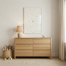 A minimalist child's room features a light-toned wooden dresser with six drawers. Above it hangs a framed vertical artwork illustrating a white bird looking up at falling pink petals. A small lamp with a textured shade and a wooden toy airplane sit on the dresser. To the left, a woven basket is filled with plush stuffed animals, with small wooden blocks on the floor nearby. The room is decorated in neutral tones with a textured rug and soft cream curtains.