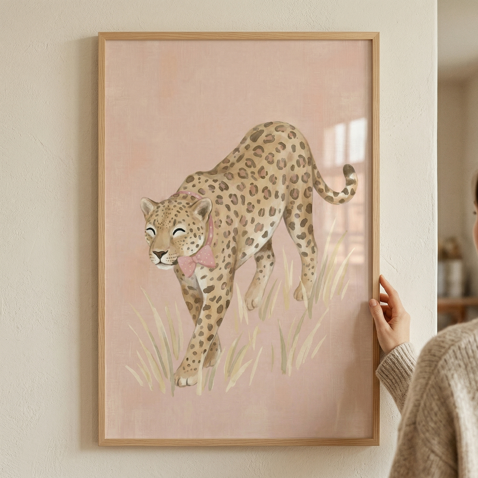 A framed piece of artwork featuring a whimsical illustration of a spotted big cat. The animal, reminiscent of a leopard, is shown in a walking pose through tall, light-colored grass. It has a gentle expression with closed eyes and is adorned with a delicate pink bow around its neck. The background is a solid, soft pink, contributing to the overall pastel and gentle aesthetic. The artwork is held in a minimalist light wood frame and is being displayed against a neutral-toned wall.