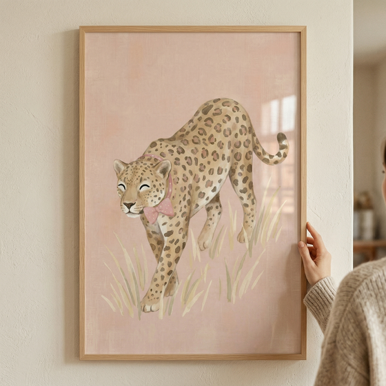 A framed piece of artwork featuring a whimsical illustration of a spotted big cat. The animal, reminiscent of a leopard, is shown in a walking pose through tall, light-colored grass. It has a gentle expression with closed eyes and is adorned with a delicate pink bow around its neck. The background is a solid, soft pink, contributing to the overall pastel and gentle aesthetic. The artwork is held in a minimalist light wood frame and is being displayed against a neutral-toned wall.