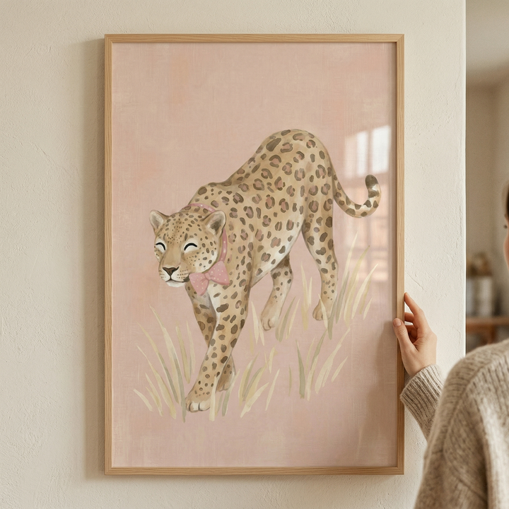 A framed piece of artwork featuring a whimsical illustration of a spotted big cat. The animal, reminiscent of a leopard, is shown in a walking pose through tall, light-colored grass. It has a gentle expression with closed eyes and is adorned with a delicate pink bow around its neck. The background is a solid, soft pink, contributing to the overall pastel and gentle aesthetic. The artwork is held in a minimalist light wood frame and is being displayed against a neutral-toned wall.