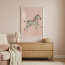A framed piece of art hangs on a soft pink wall in a serene nursery setting. The artwork features a whimsical, watercolor-style illustration of a young zebra leaping through the air, wearing a delicate pink ribbon bow around its neck. Below the frame sits a light-toned wooden dresser with two drawers, topped with a small cream-colored lamp and a round woven basket. To the left, a plush pink armchair is partially visible, draped with a cozy, cream-colored knitted throw blanket.