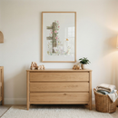 A minimalist nursery or bedroom scene features a light-toned wooden dresser with three wide drawers. Centered above it, a wooden-framed piece of art depicts a weathered fence post entwined with climbing pink roses and delicate wildflowers, standing next to a serene white goose. Atop the dresser are natural wood toy blocks, a toy car, and a small green plant in a white ceramic pot. A woven wicker basket filled with folded blankets sits on the light-colored, textured rug beside the dresser.