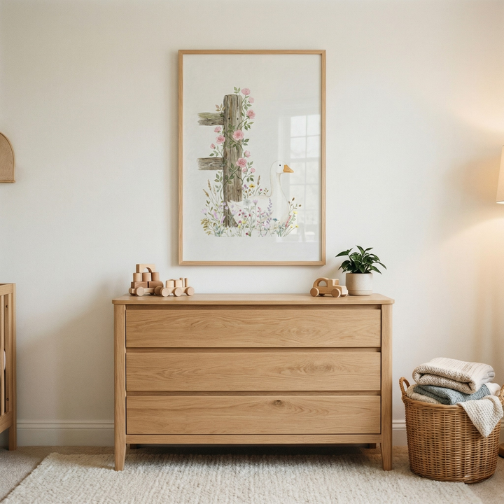 A minimalist nursery or bedroom scene features a light-toned wooden dresser with three wide drawers. Centered above it, a wooden-framed piece of art depicts a weathered fence post entwined with climbing pink roses and delicate wildflowers, standing next to a serene white goose. Atop the dresser are natural wood toy blocks, a toy car, and a small green plant in a white ceramic pot. A woven wicker basket filled with folded blankets sits on the light-colored, textured rug beside the dresser.