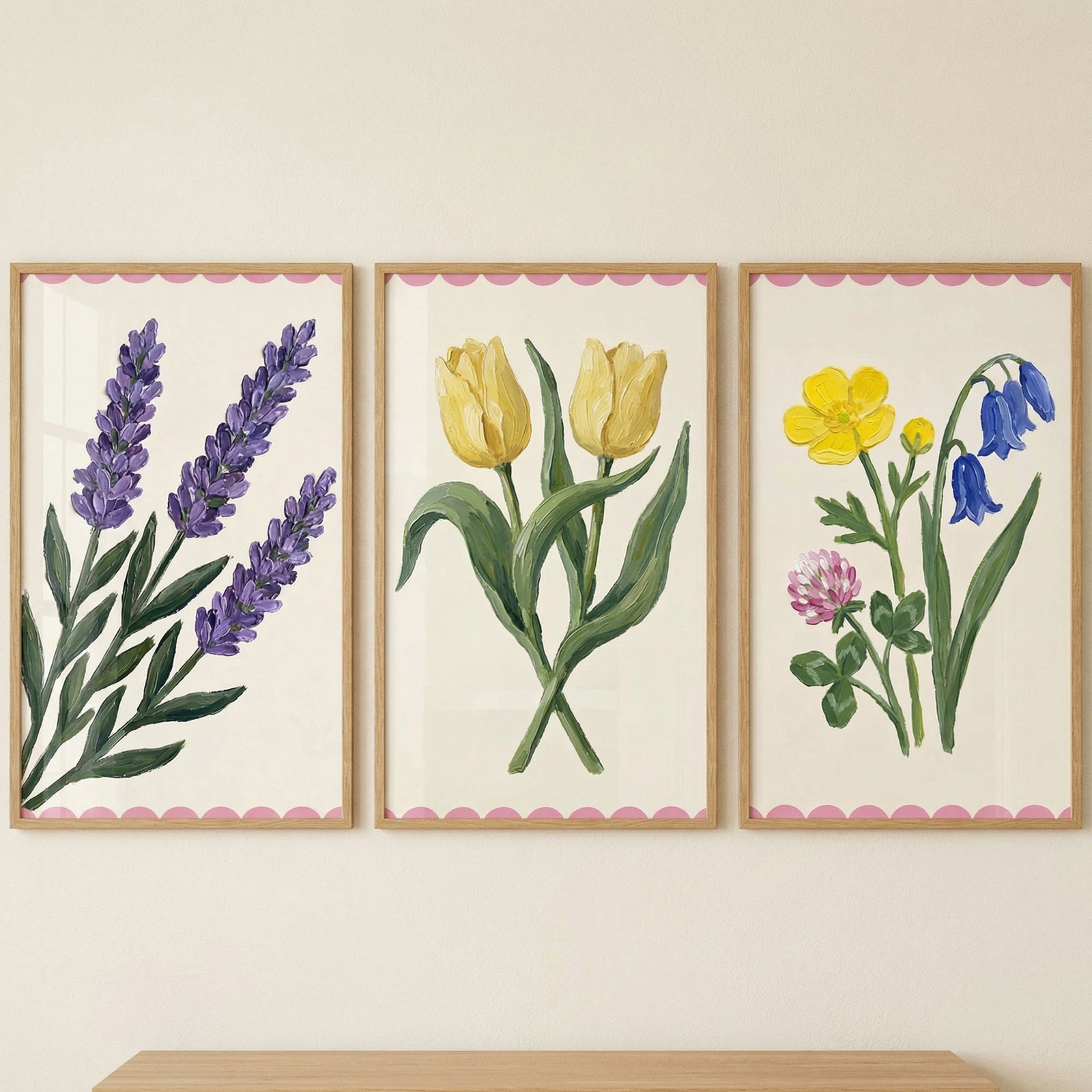 Three framed botanical prints on a wall featuring purple lavender on the left two yellow tulips in the center and a mix of small wildflowers on the right each painted in a soft hand painted style with a pink scalloped border.