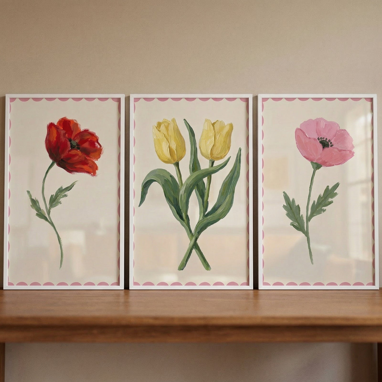 Three framed floral paintings with pink scalloped borders displayed side by side on a wooden surface featuring a red poppy yellow tulips and a soft pink bloom creating a warm balanced botanical arrangement.