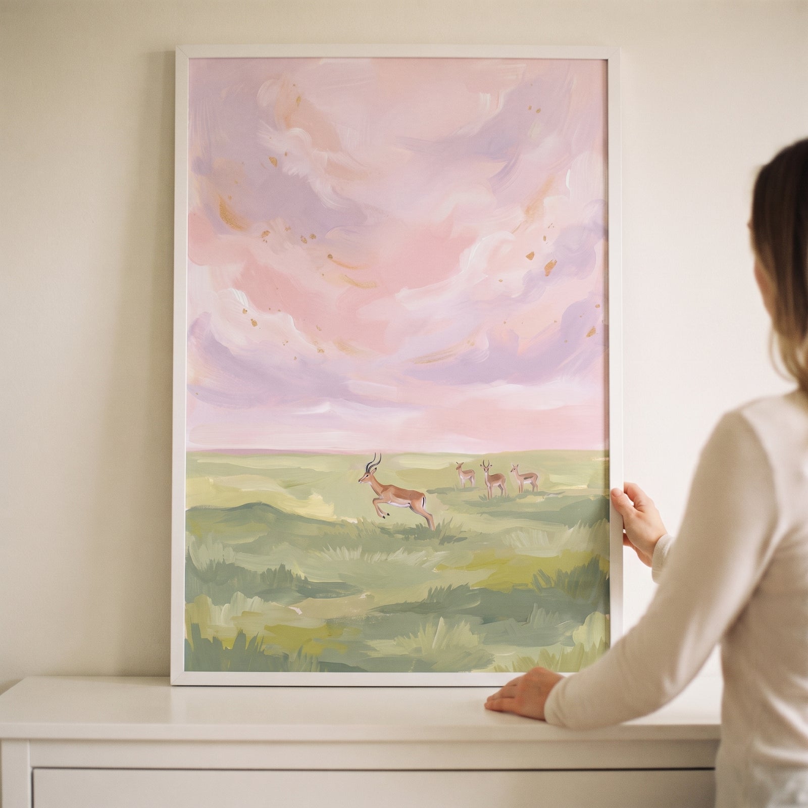A person holds pink sky leaping gazelle animal wall art in a white frame above a white dresser.