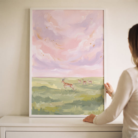 A person holds pink sky leaping gazelle animal wall art in a white frame above a white dresser.