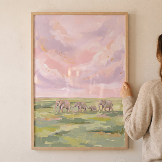A person holds a framed pink sky walking elephants animal wall art showing four elephants in a green field under soft clouds.