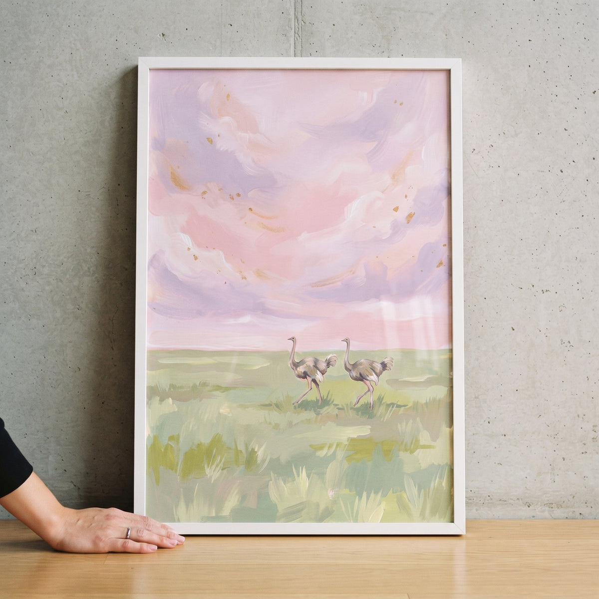 Pink sky walking ostrich animal wall art depicts two birds in a grassy field under soft pastel clouds in a white frame.