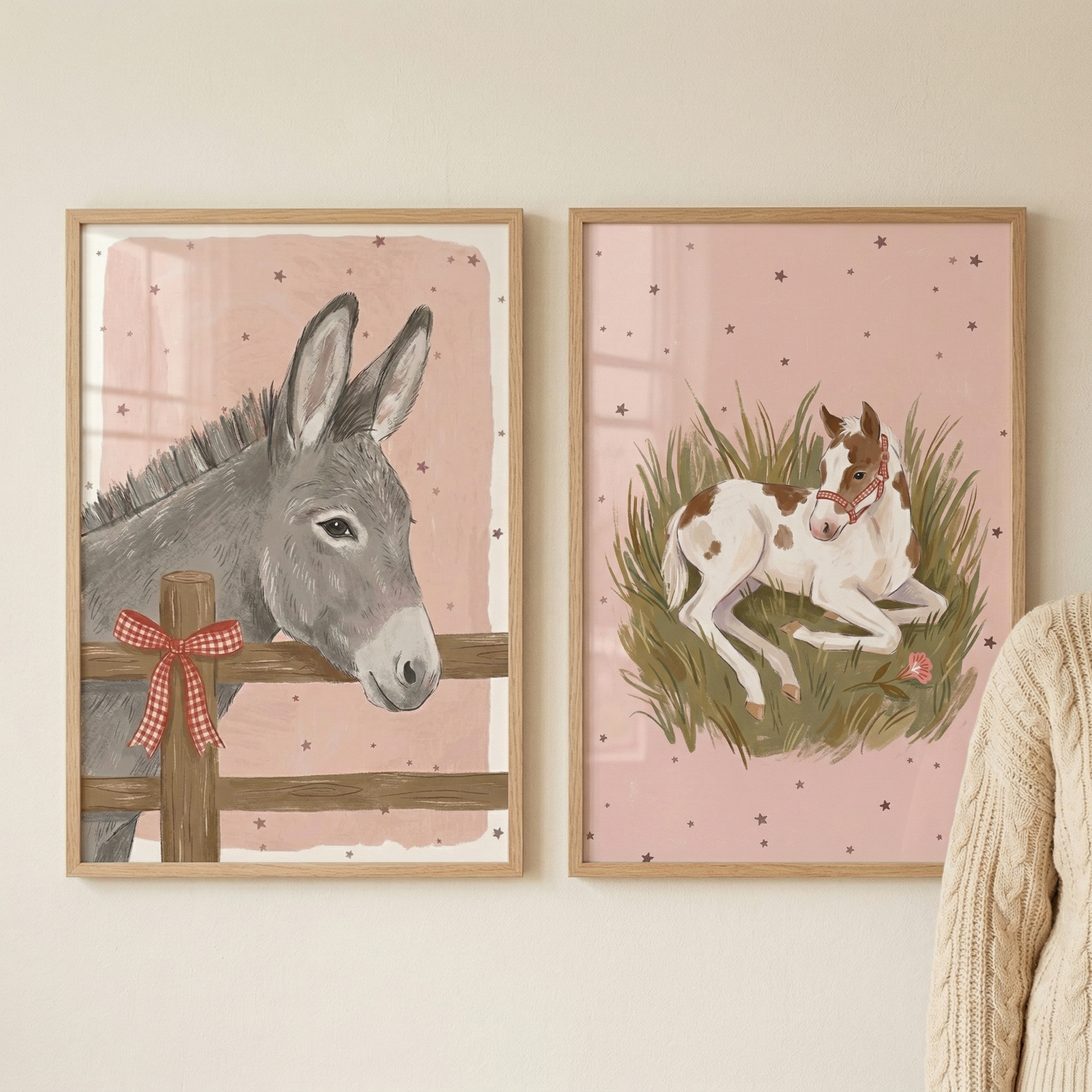 Two framed illustrations hang side-by-side on a neutral-toned wall. The left artwork features a grey donkey looking over a simple wooden fence with a red and white checkered bow tied to a post. The right artwork shows a brown and white spotted foal resting in green grass, wearing a delicate red halter. Both scenes share a soft pink background decorated with tiny, dark stars and are encased in light-toned wooden frames. A portion of a cream cable-knit sweater is visible on the right side.