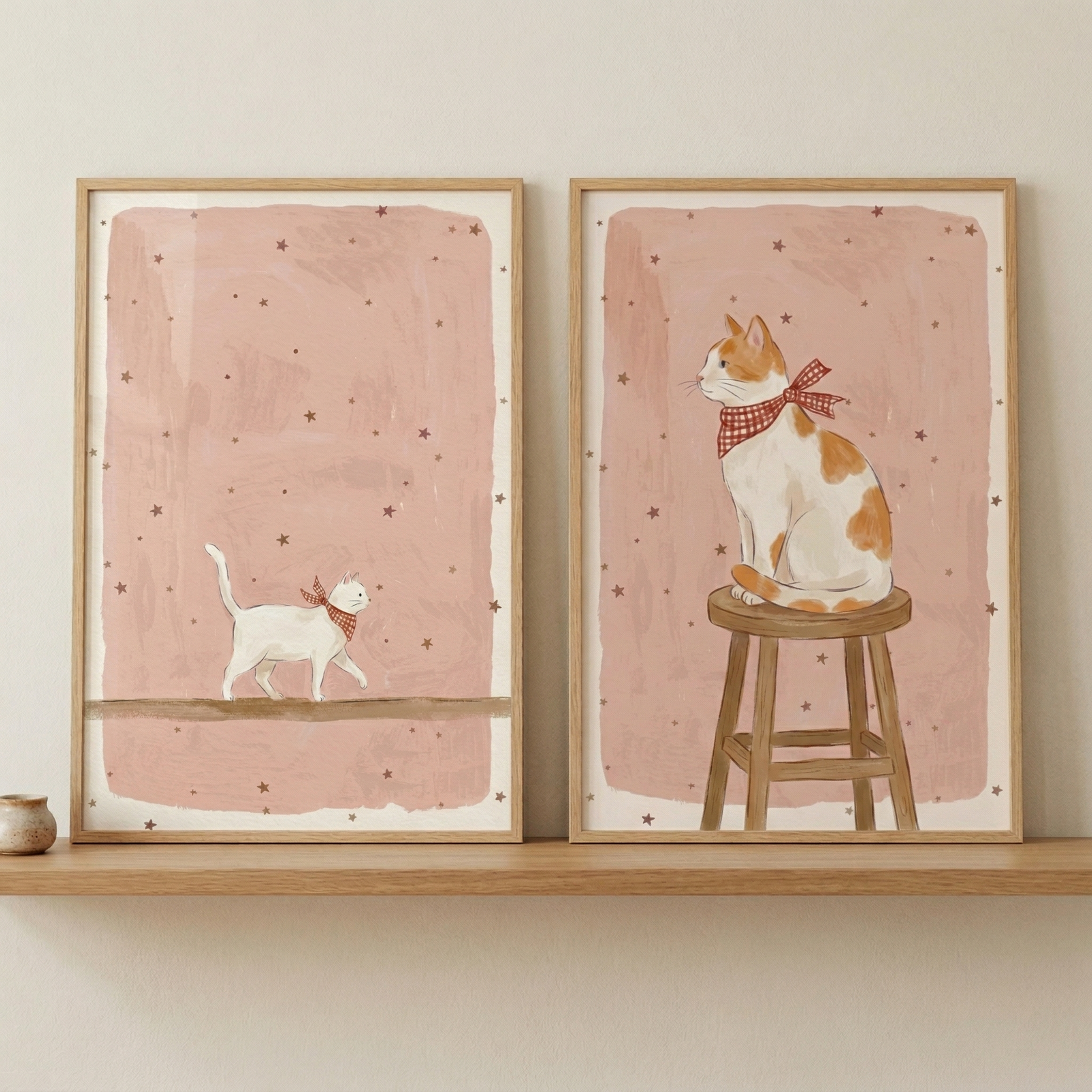 Two vertical wooden frames sit side-by-side on a light-colored shelf against a neutral wall. Each frame contains a whimsical illustration with a dusty pink textured background scattered with tiny dark stars. 

The left piece shows a small white cat with ginger spots walking across a thin horizontal line, wearing a red-and-white checkered scarf. 