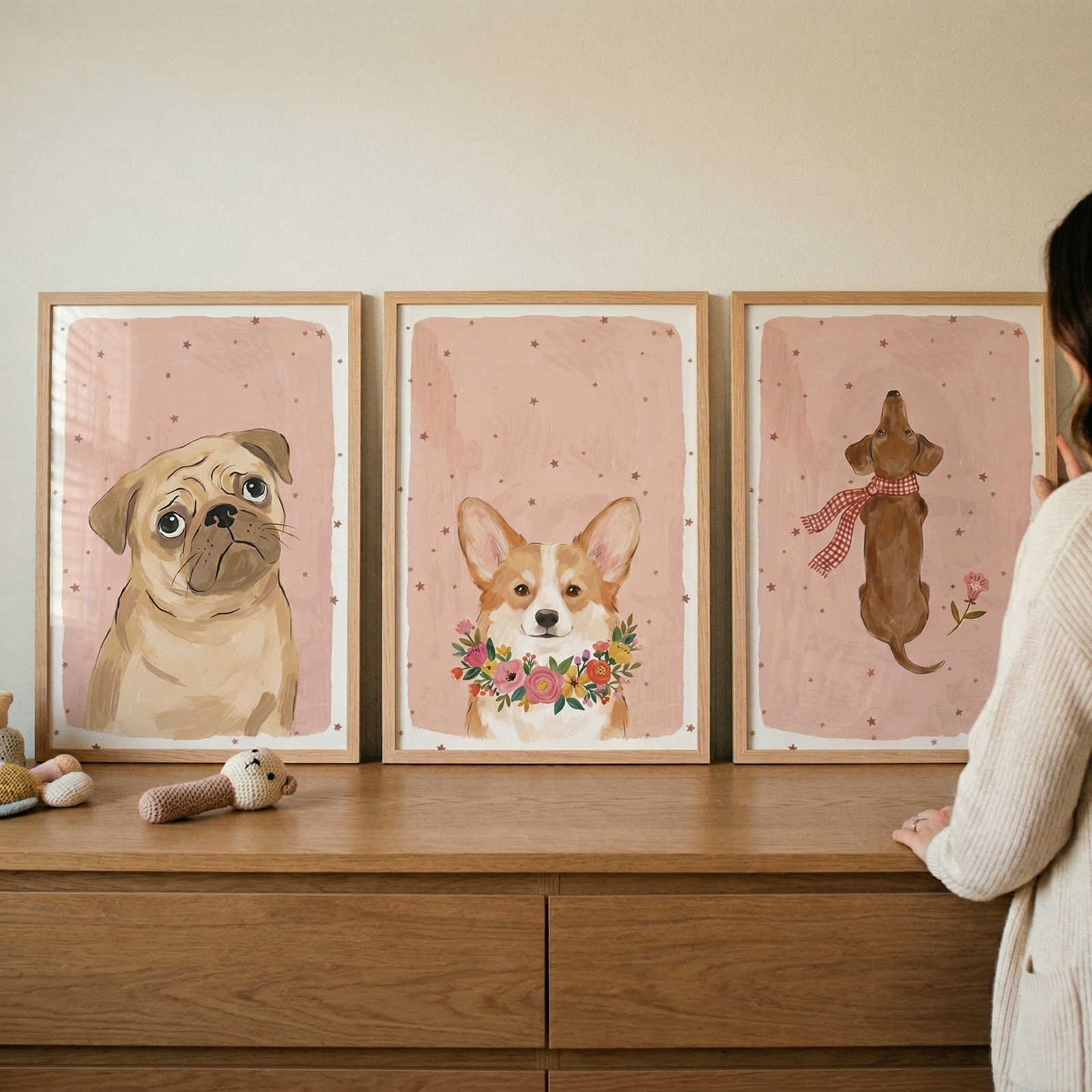 Three wooden-framed illustrations of dogs are displayed on a light oak dresser. Each piece features a different dog against a soft pink background dotted with small, dark stars.