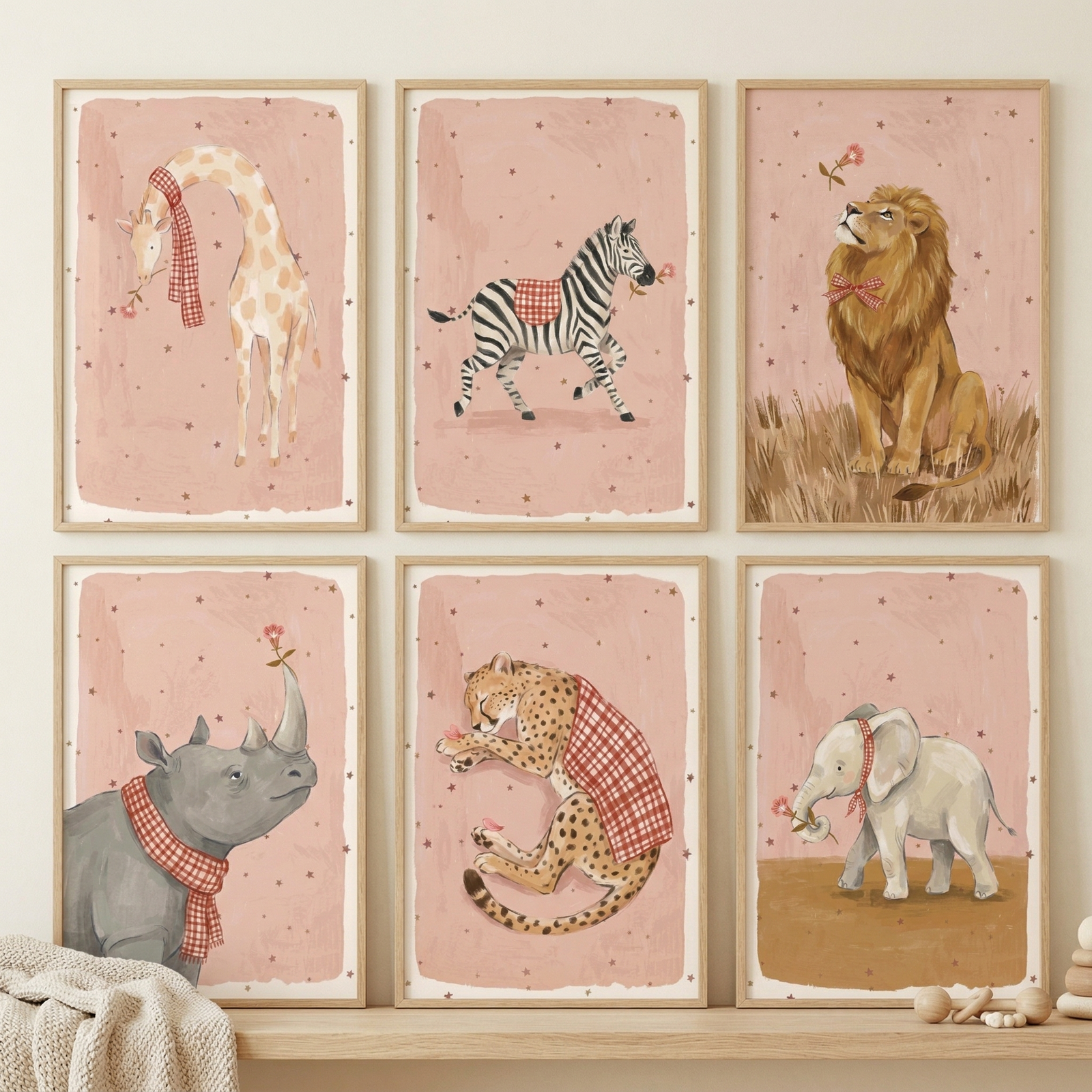 A set of six framed illustrations of safari animals is displayed in two rows of three on a light wood shelf. Each animal is depicted in a whimsical style with soft colors and a recurring red-and-white checkered fabric theme. The backgrounds are a gentle dusty rose color accented with small, dark star-like dots.
