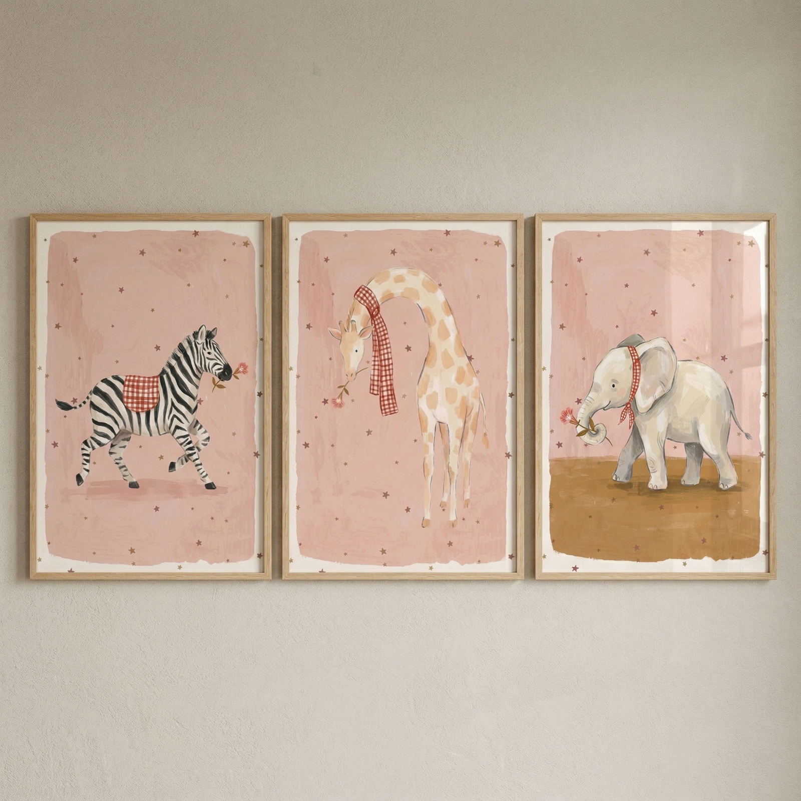 A set of three framed artworks hangs side-by-side on a neutral wall. Each piece features a whimsical animal illustrated in a soft watercolor style against a dusty pink background speckled with small stars. In the left frame, a zebra wears a red and white checkered blanket. 