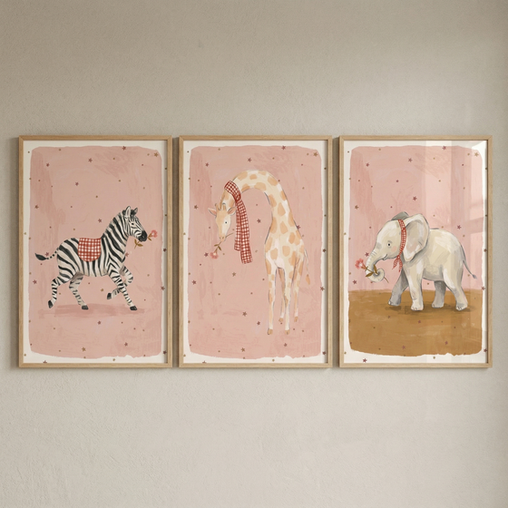 A set of three framed artworks hangs side-by-side on a neutral wall. Each piece features a whimsical animal illustrated in a soft watercolor style against a dusty pink background speckled with small stars. In the left frame, a zebra wears a red and white checkered blanket. 