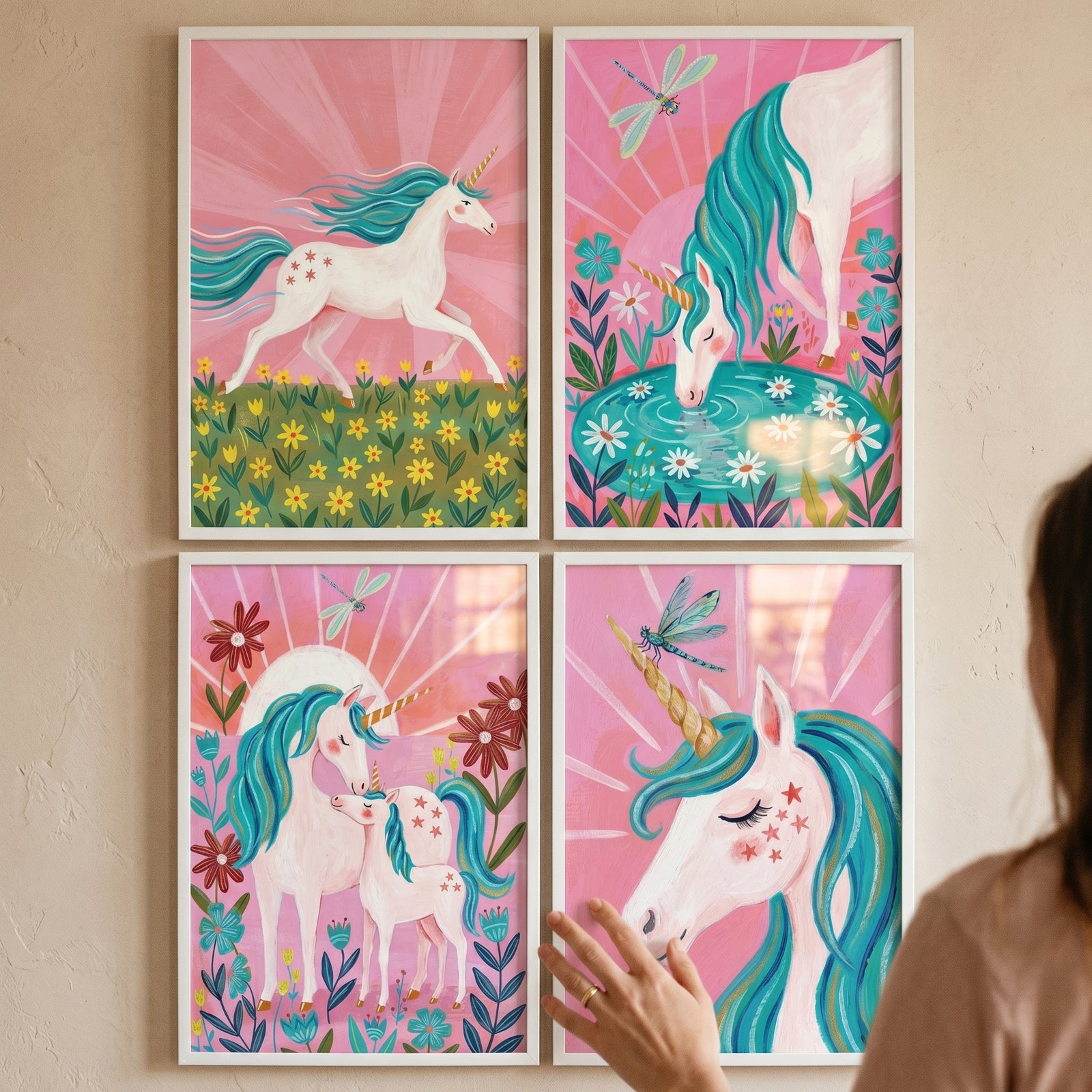A pink sunburst whimsical unicorn wall art set of 4 framed illustrations displayed on a wall with a hand touching them.