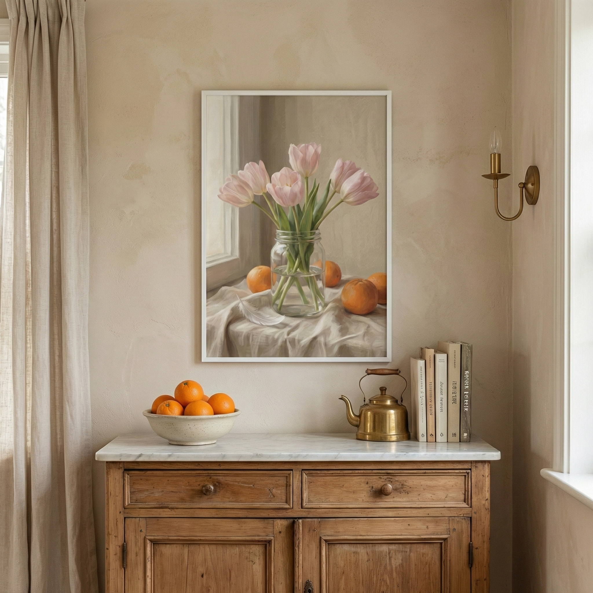 A cozy corner with a wooden dresser topped with a white marble surface, featuring a bowl of oranges, a brass teapot, and a stack of books. Above, a framed painting depicts pink tulips in a jar surrounded by oranges on a draped cloth, all set against a soft beige wall with a single brass sconce.