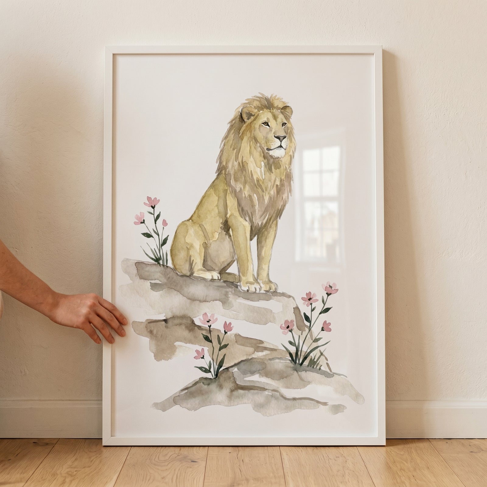 Pink wildflower lion wall art shows a watercolor lion sitting on rocks among pink blossoms in a simple white frame.