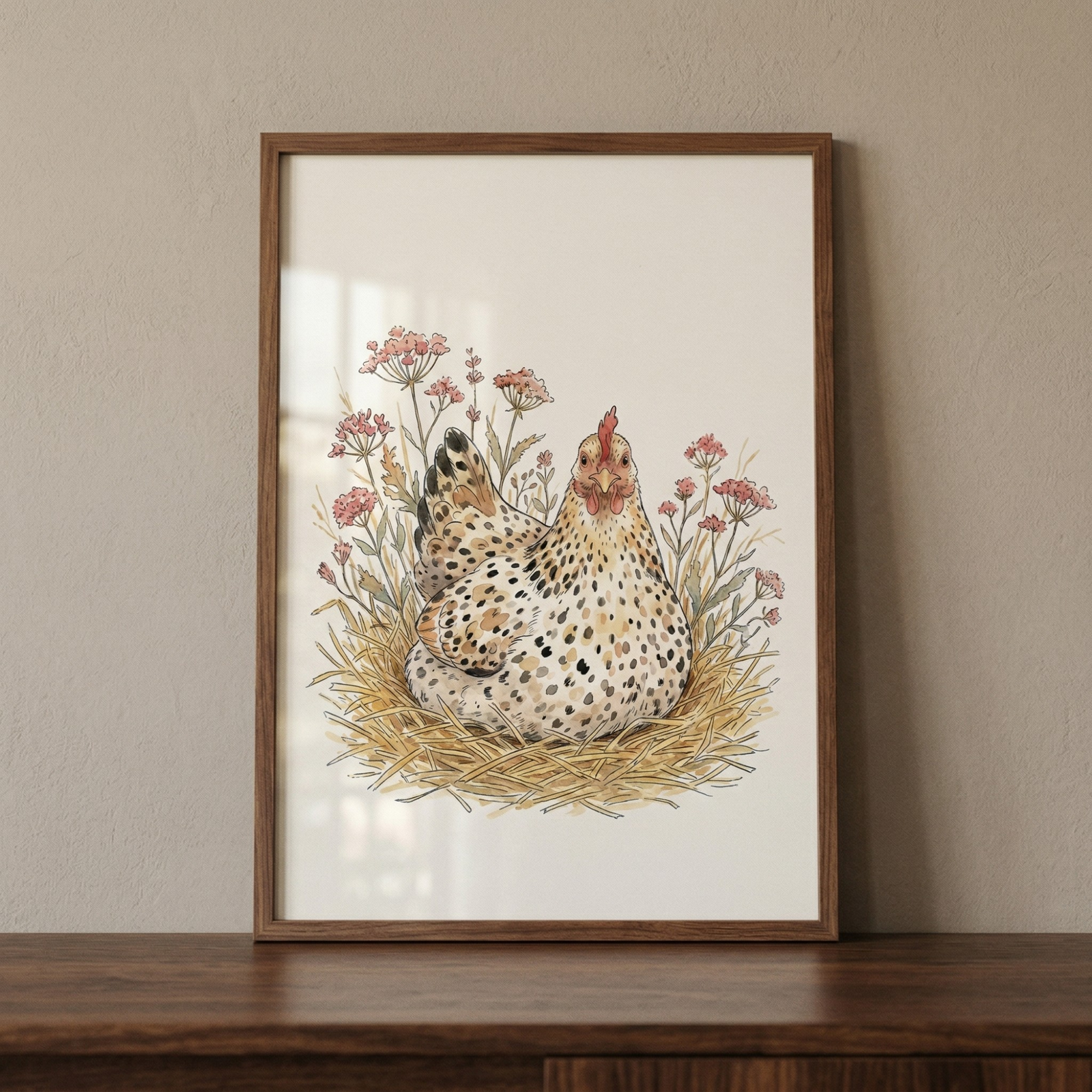 A framed illustration of a speckled hen resting in a straw nest, surrounded by soft pink wildflowers. The artwork is done in a delicate, watercolor style with earth tones and fine lines. The picture is held in a simple brown wooden frame, which sits on a dark wood surface against a plain, warm-toned wall.