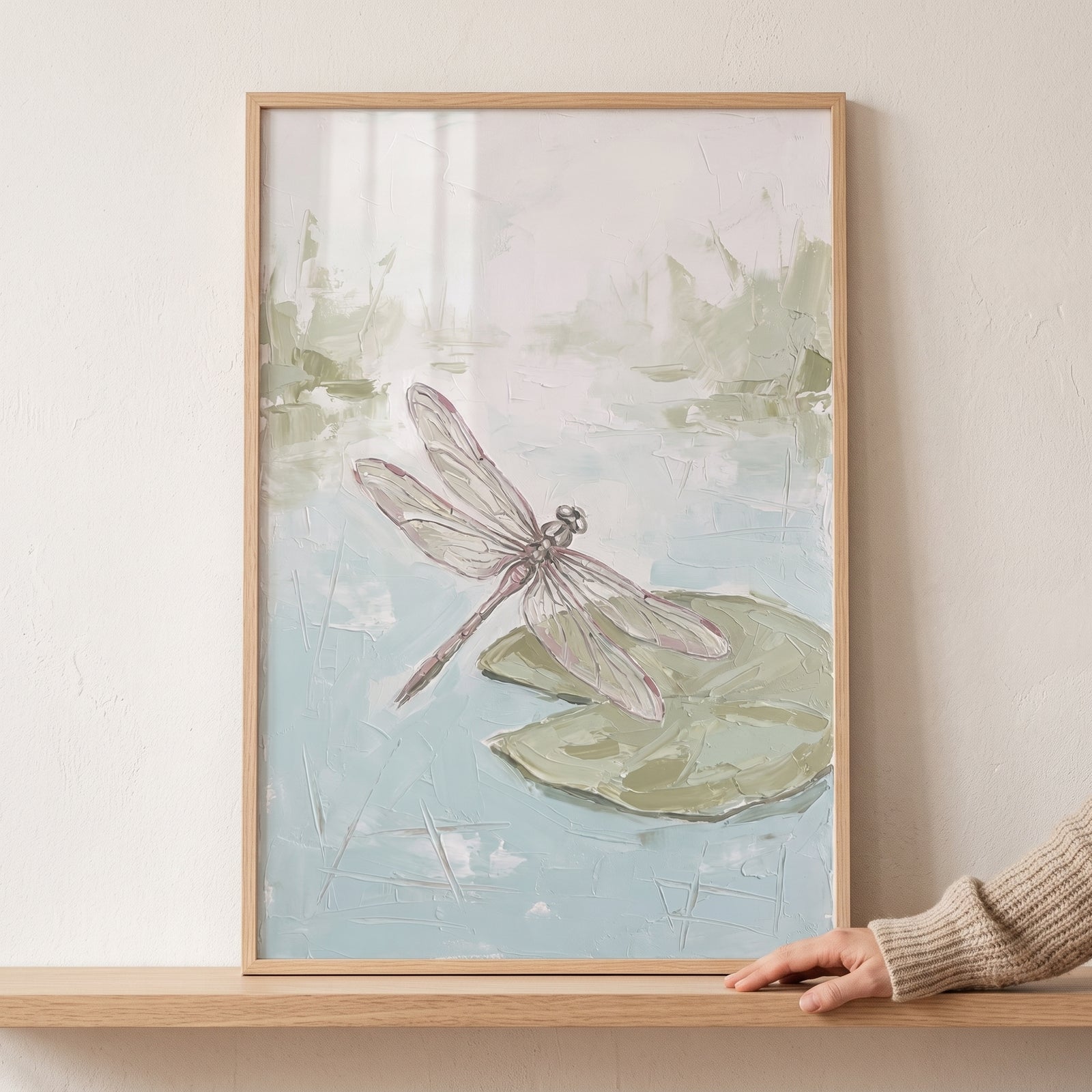 Pink wing textured dragonfly pond wall art in a light wood frame rests on a shelf next to a hand.