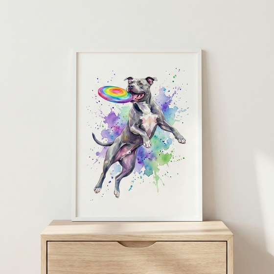A watercolor illustration of a grey and white short-haired dog leaping through the air. The dog is shown in a dynamic pose, reaching for a colorful, rainbow-patterned flying disc. The background is filled with expressive splashes and drips of purple, blue, and green watercolor. The artwork is housed in a clean white frame and displayed on a light-toned wooden dresser against a plain white wall.