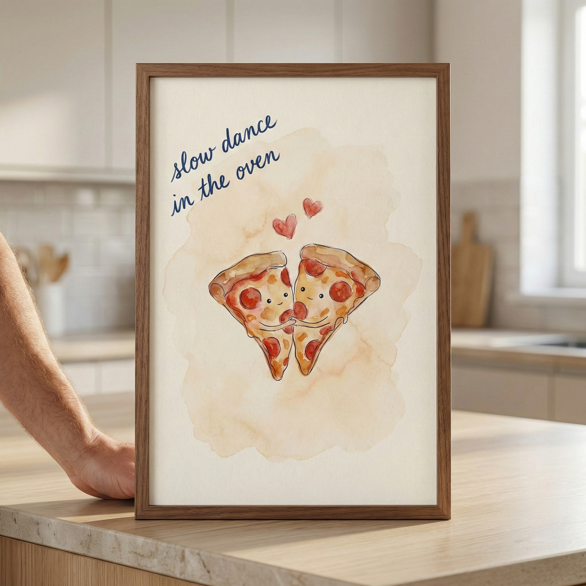 A framed illustration of two smiling pizza slices holding hands as if they are dancing. Above the slices are two small red hearts, all set against a soft, light-orange watercolor-style wash. In the top-left corner, the words "slow dance in the oven" are written in a blue cursive font. The dark wood-framed piece sits on a wooden kitchen counter.