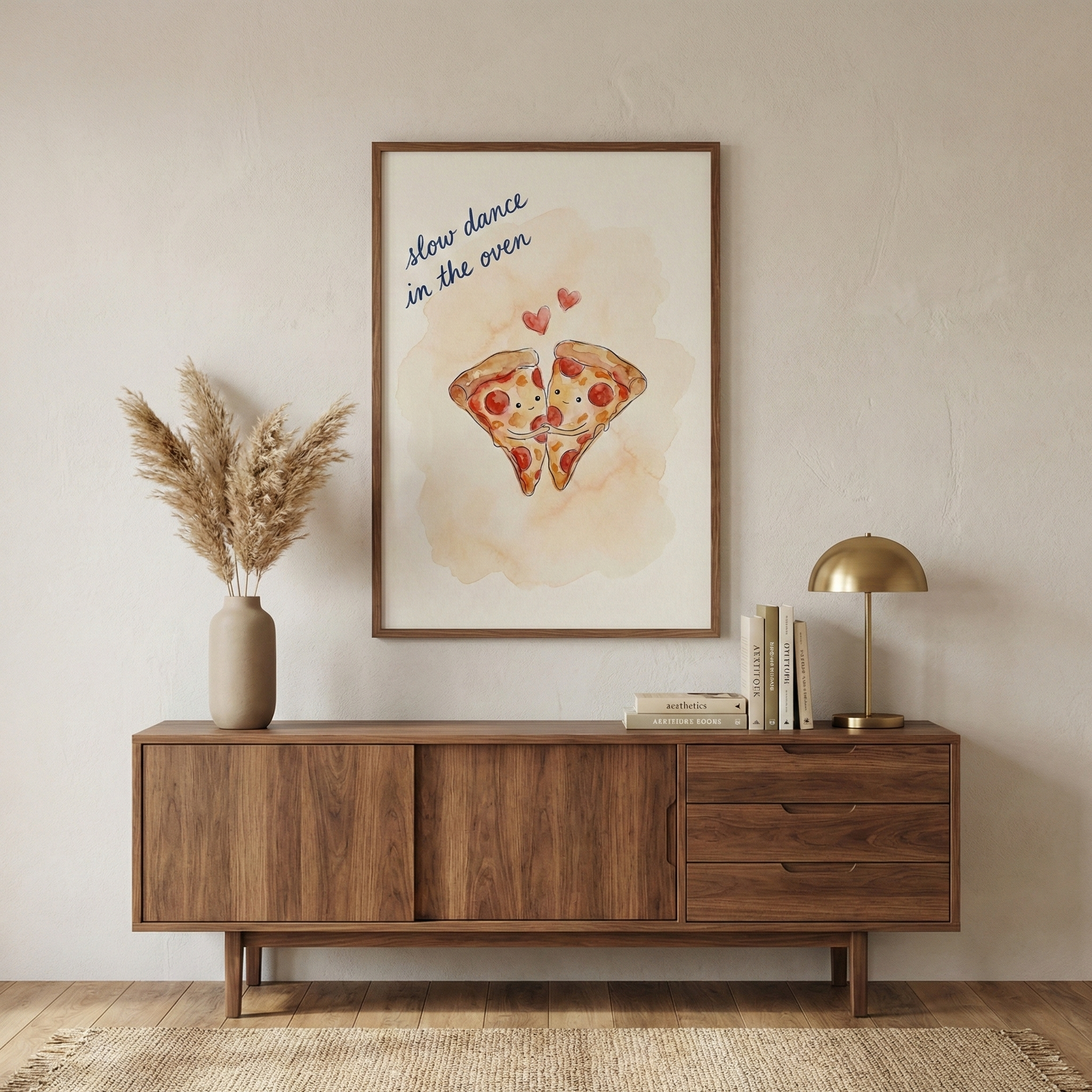 A framed piece of wall art hangs on a light-colored, textured wall above a wooden mid-century modern sideboard. The art depicts two cartoon pizza slices with small faces and arms, appearing to hug or dance together against a soft watercolor wash. Above them, the text "slow dance in the oven" is written in blue, accompanied by small red heart shapes. On the sideboard, a beige ceramic vase holds dried feathery grasses to the left, while a small stack of books and a gold-finished dome lamp sit to the right. 