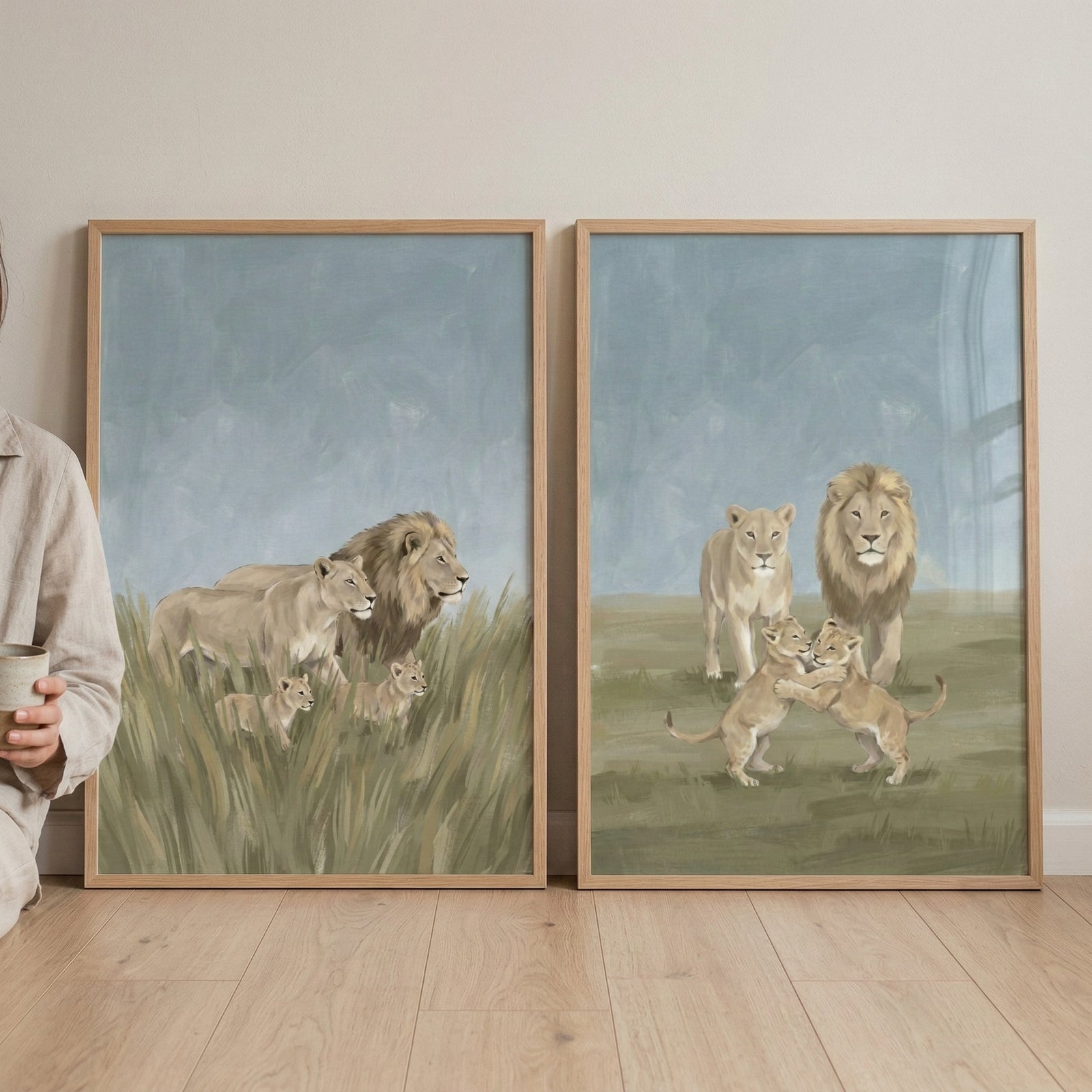 Two framed vertical illustrations featuring lion families are displayed side-by-side, leaning against a neutral-toned wall on a light wood floor. The piece on the left depicts a male lion, a lioness, and two small cubs amidst tall, golden-green grass. The piece on the right shows the same family on a shorter grassy field, with the two cubs playfully wrestling in the foreground while the adults watch from behind. Both illustrations use a soft, textured style with a palette of muted earthy browns and greens u