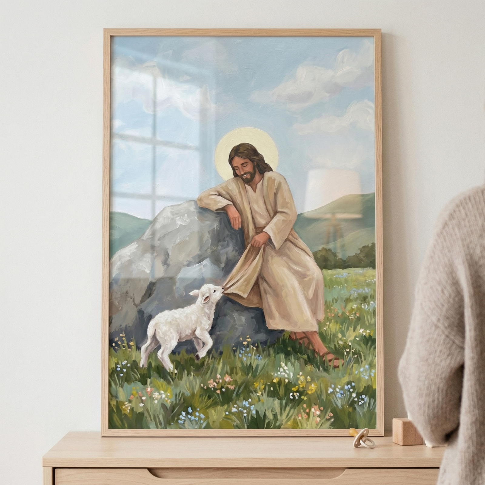 A framed vertical artwork depicts Jesus Christ sitting on a large grey rock in a lush meadow filled with wildflowers. He wears a light tan robe and has a golden halo around his head. A small white lamb stands at his feet, playfully tugging on the hem of his robe. Jesus looks down at the lamb with a gentle smile. The background features soft rolling hills under a light blue sky with wispy white clouds. The art is held in a light wood frame and placed on a wooden dresser.
