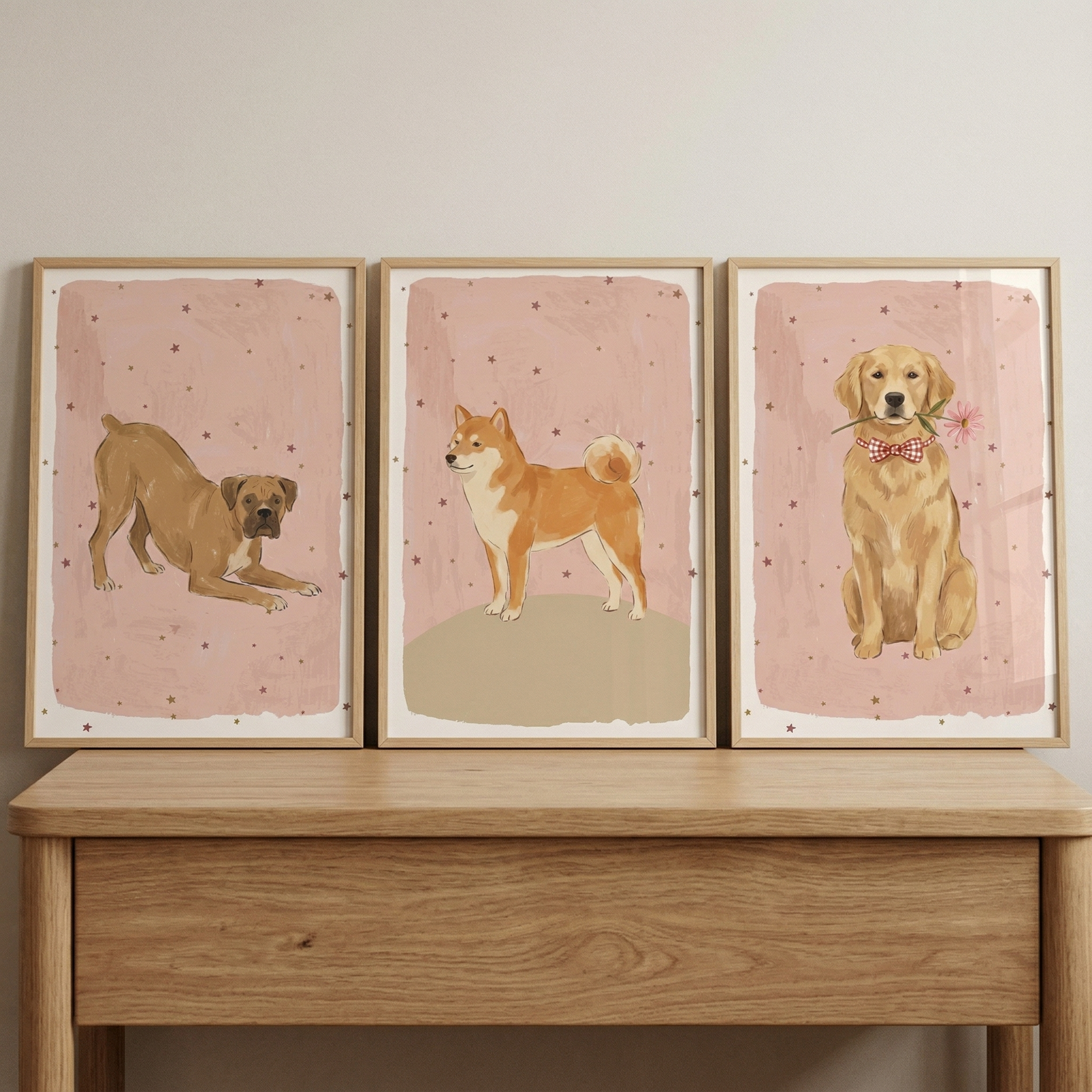 Three wooden-framed illustrations of dogs are displayed side-by-side on a light wood console table against a neutral wall. Each artwork features a different dog breed set against a dusty pink background speckled with small brown stars. The left illustration shows a tan-colored dog in a playful pose. 