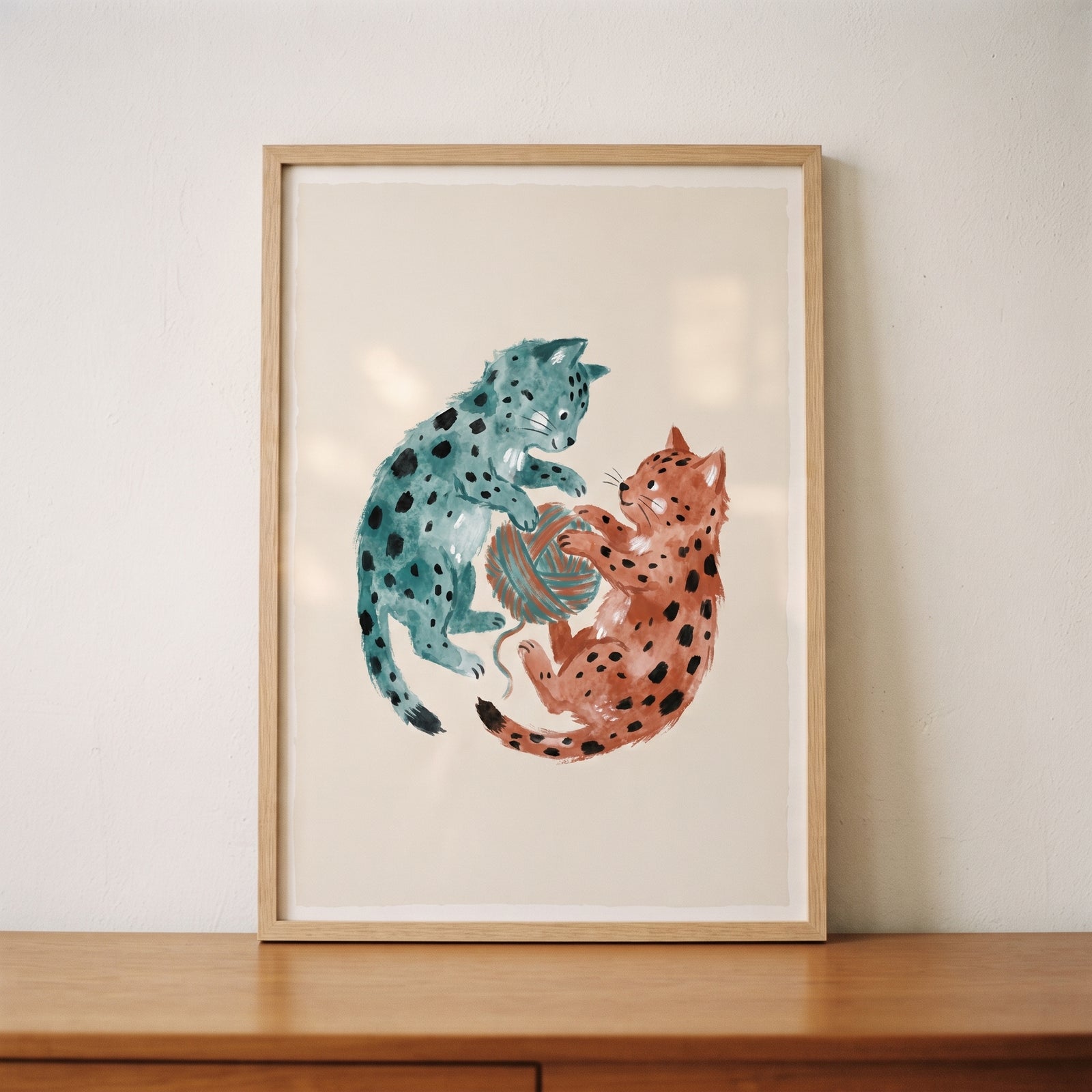 Teal and orange playing yarn spotted kittens wall art in a light wood frame on a table against a white wall.