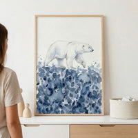 Framed watercolor painting of a polar bear walking across an abstract patch of icy blue splashes, suggesting snow or frozen terrain. The soft, wintry tones and minimalist background create a calm and cozy atmosphere.