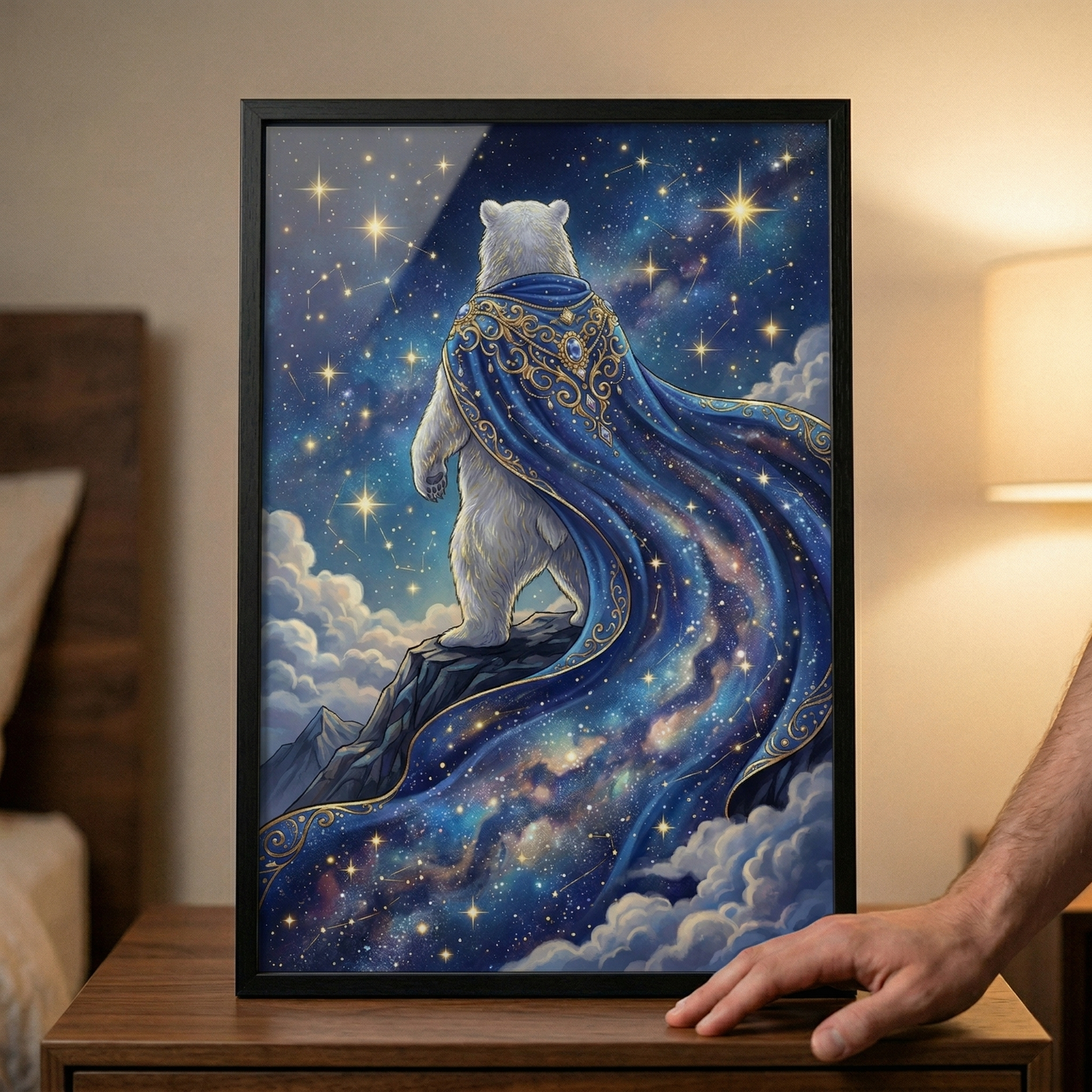 A framed digital illustration shows a white polar bear standing on a rocky mountain ledge, seen from behind. It wears a majestic, flowing blue cape with intricate gold filigree around the neck. The cape’s fabric merges into a vast, swirling galaxy filled with stars and nebulae. The bear looks out over soft clouds into a deep blue sky crowded with glowing stars. The artwork is held in a black frame, resting on a wooden table in a dimly lit room next to a soft light source.