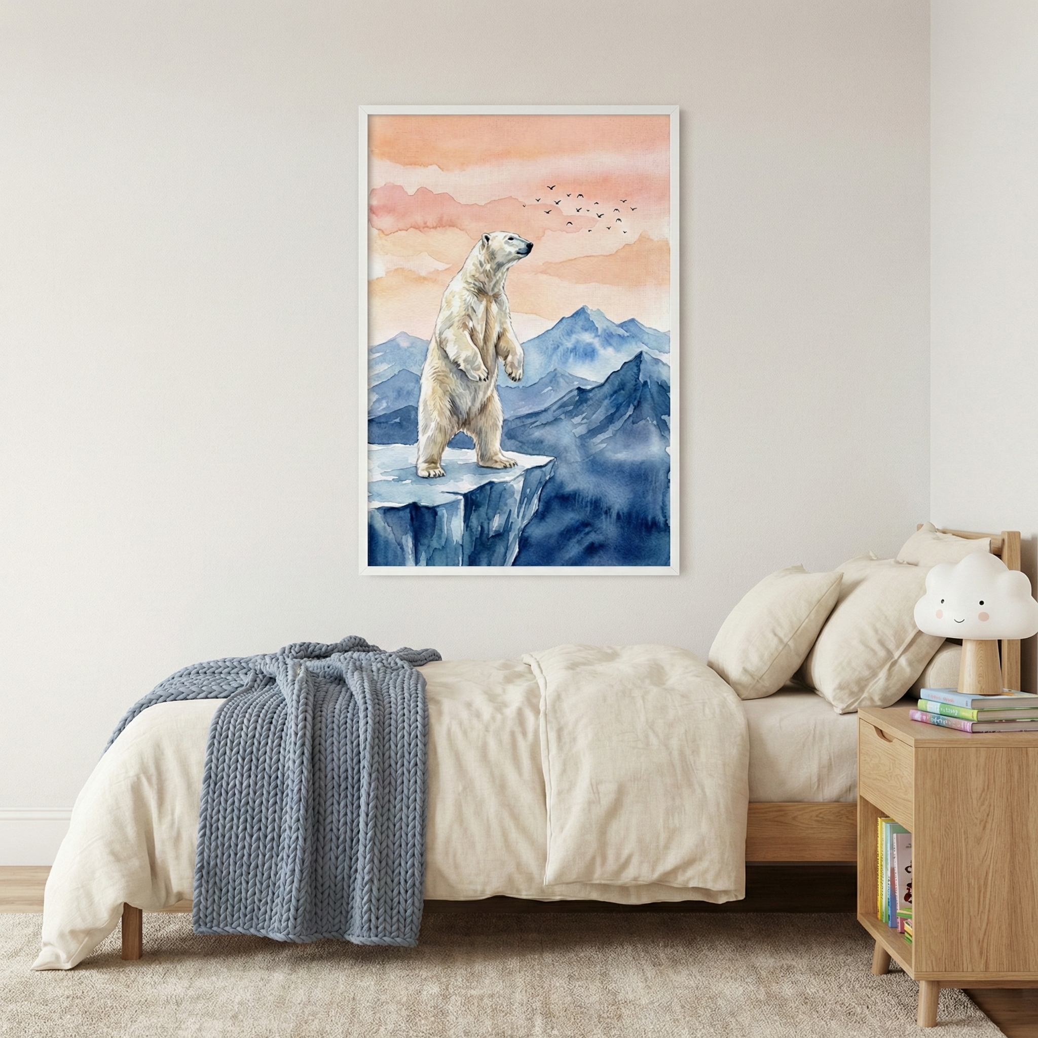 A minimalist child's bedroom features a large, framed rectangular artwork centered on a white wall. The illustration depicts a polar bear standing on its hind legs on a snowy cliff, overlooking a range of blue mountains under a soft peach and orange sky with small birds in the distance. Below the art, a single bed is made with light cream linens and a chunky blue knit blanket. To the right, a wooden nightstand holds a small, smiling cloud lamp and several books. A neutral rug covers the floor.