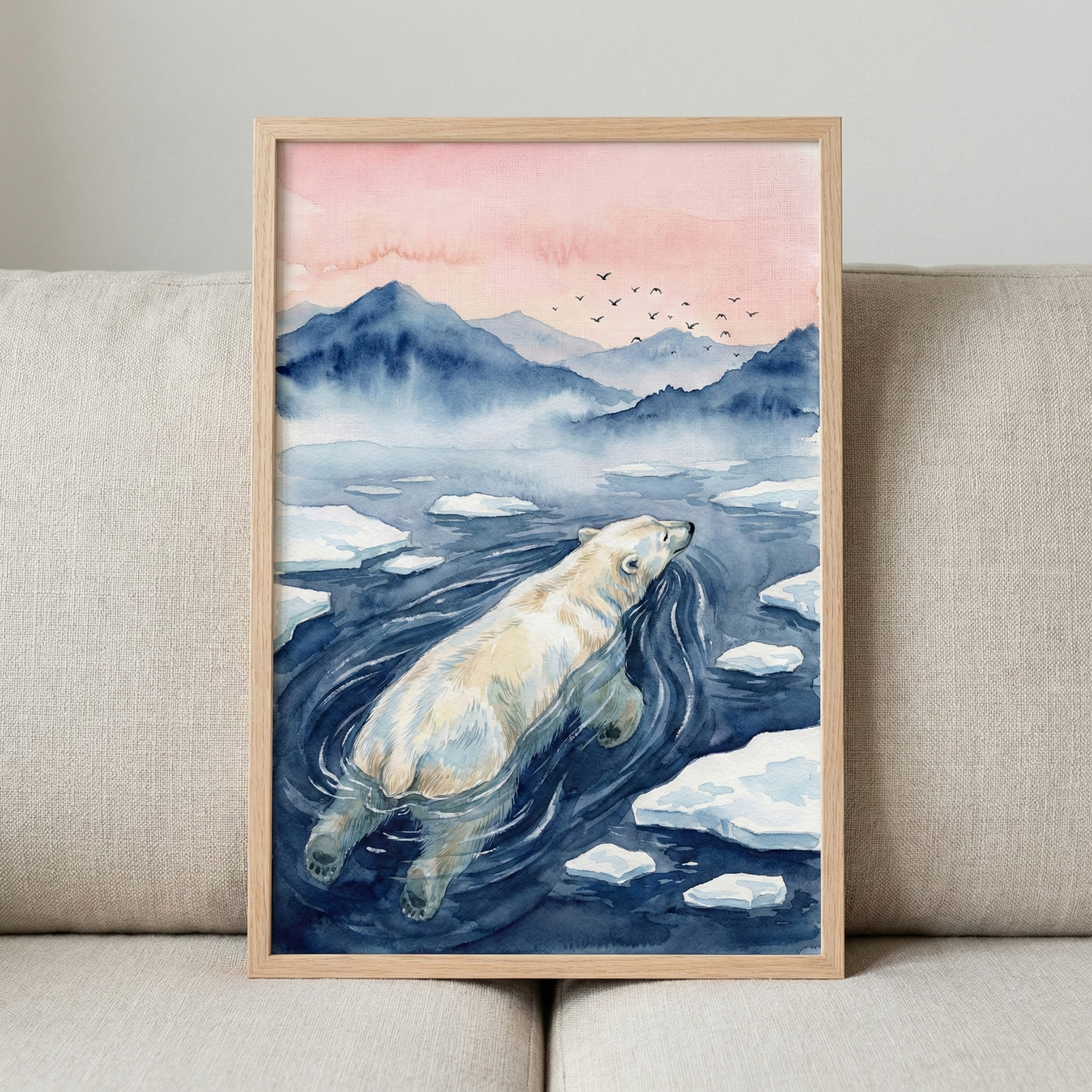 A framed piece of watercolor-style artwork sits propped against a neutral, textured sofa. The illustration depicts a large polar bear swimming through deep blue water, past several white ice floes. In the background, misty blue mountain peaks rise toward a soft, pale pink and orange sunset sky. A small flock of birds flies in the distance. The art is enclosed in a simple, light-toned wooden frame, creating a serene and natural scene.