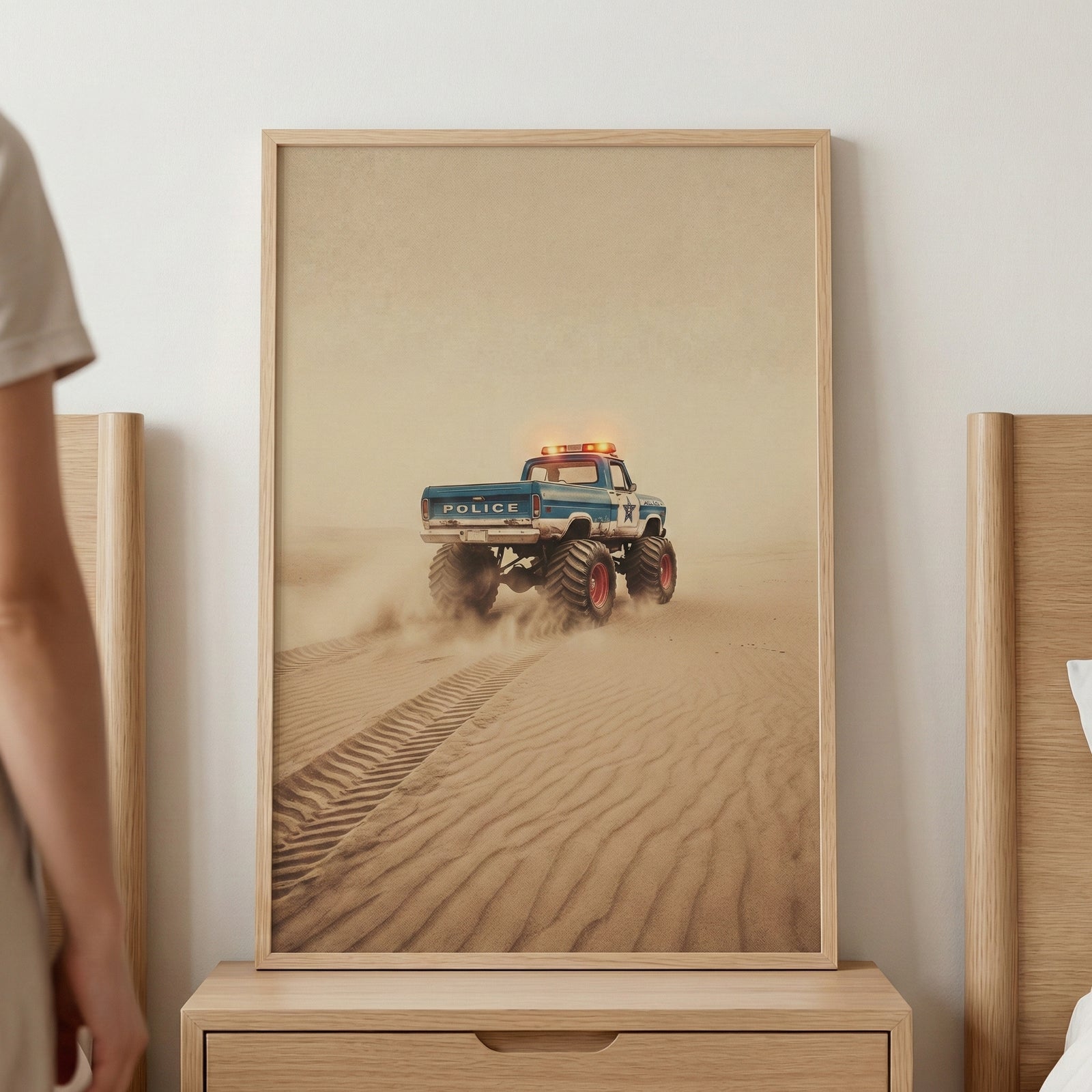A framed vertical artwork sits on a light wood nightstand, depicting a large blue and white off-road patrol truck with oversized tires driving across a vast desert. The vehicle, equipped with glowing amber lights on its roof, kicks up dust as it traverses the rippling sand dunes, leaving deep tire tracks in its wake. The scene is captured in a soft, hazy light with a warm, sandy color palette.