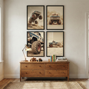 Four framed art pieces depicting monster trucks are arranged in a square on a white wall. The top left image shows a blue monster truck jumping off a dirt hill against a dusty sky. To its right, a blue monster truck is shown from the front, kicking up a cloud of dust. Below the first image, a close-up shot focuses on the massive, treaded tire of a blue and white monster truck. 