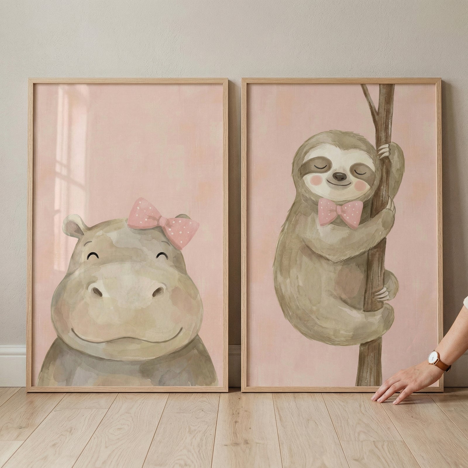 Two framed pieces of nursery artwork are displayed side-by-side on a light-colored wooden floor against a neutral wall. Both pieces feature cute, smiling cartoon animals illustrated in a soft, watercolor-like style against a blush pink background.
