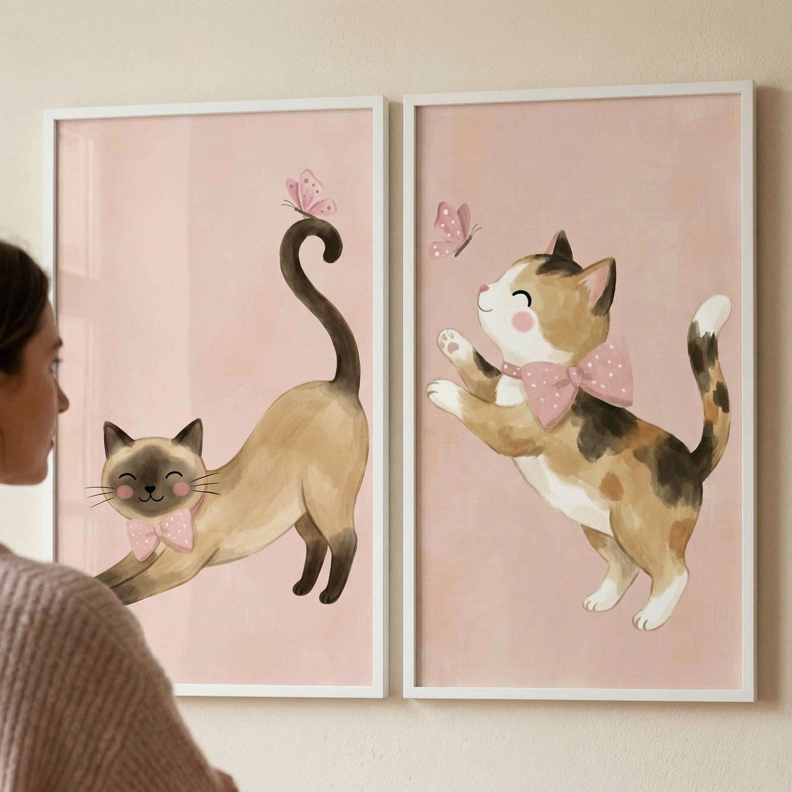 Two vertically oriented, white-framed illustrations are mounted side-by-side on a light-colored wall. Both pieces share a soft, solid light pink background. 

The illustration on the left depicts a slender, tan-colored cat with dark brown features on its face, ears, and paws. The cat is shown in a playful stretching pose, wearing a simple pink bow tie. Its long, dark tail curves upward, and a small pink butterfly rests on its tip. 