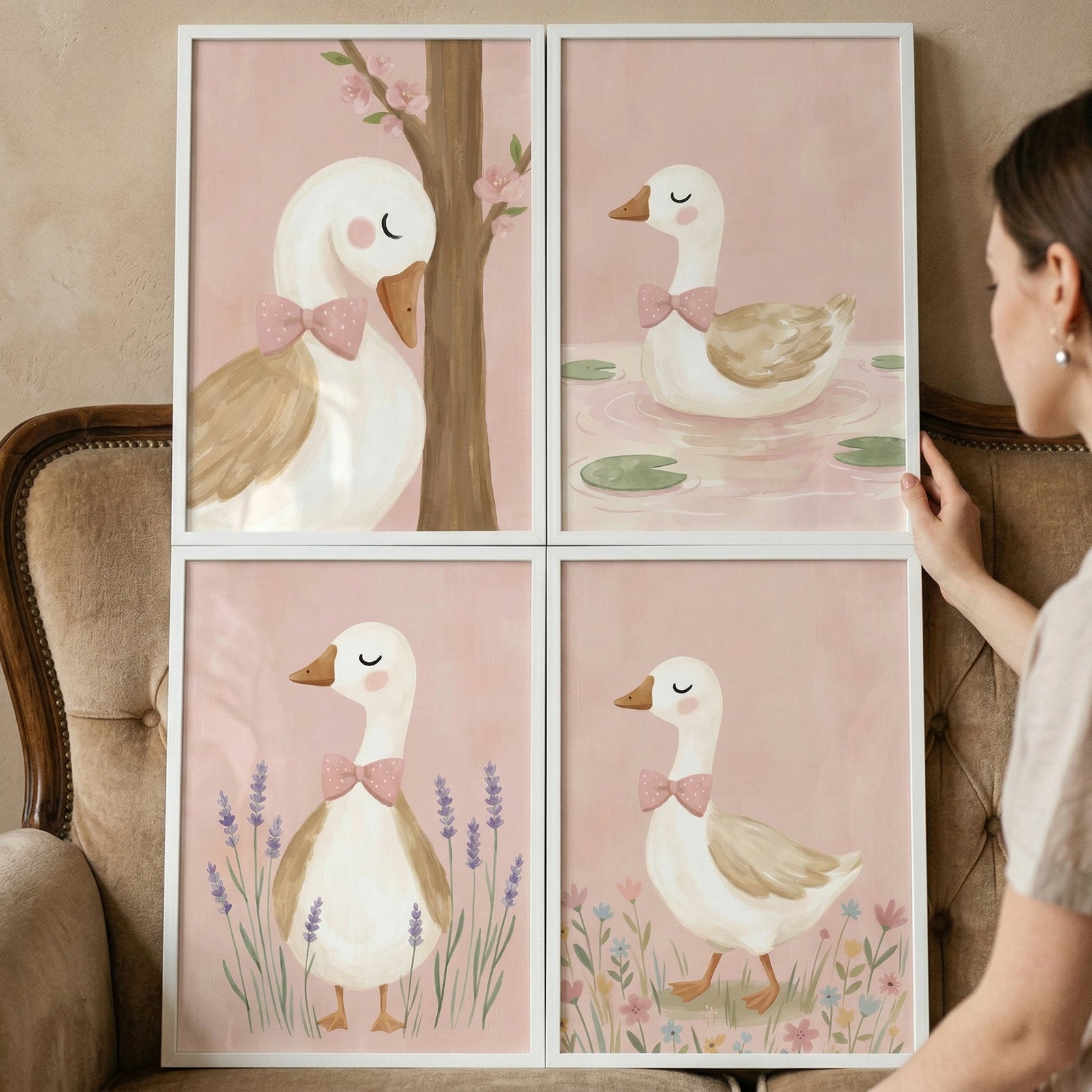 Four framed pastel duck illustrations arranged in a grid are being styled against a wall beside an upholstered chair. Each print shows a white duck with a pink bow tie in different scenes including by a tree, in water with lily pads, among lavender, and in a field of flowers, creating a soft cohesive nursery gallery wall.
