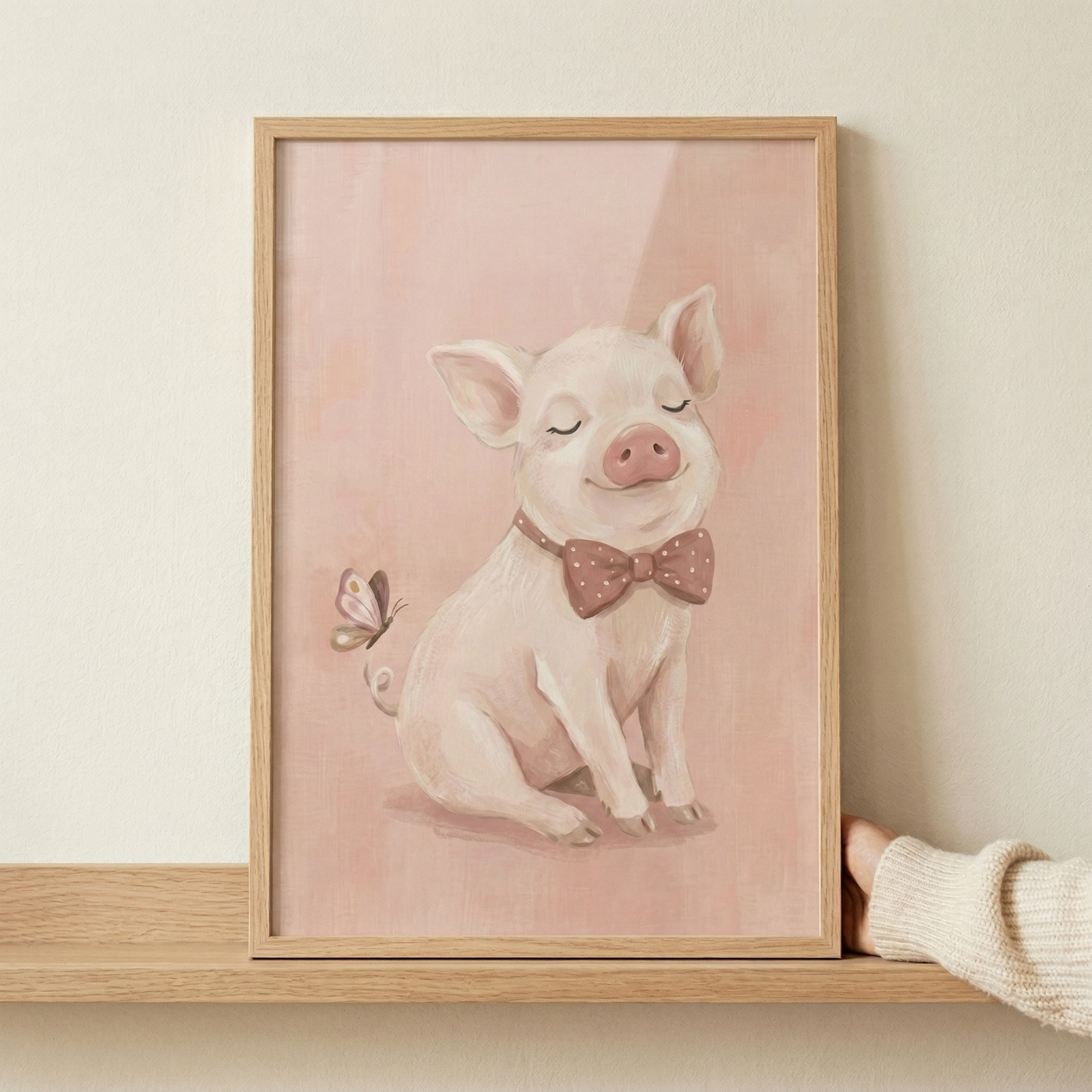 A framed illustration features a charming pink piglet sitting with a peaceful expression and its eyes closed. The piglet is adorned with a reddish-brown bow tie covered in tiny white polka dots. A delicate butterfly with brown and cream wings flutters near its curly tail. The artwork is set against a soft, textured pink background. The piece is encased in a simple light wood frame, which sits on a wooden shelf. A hand wearing a white knitted sweater is seen holding the side of the frame.