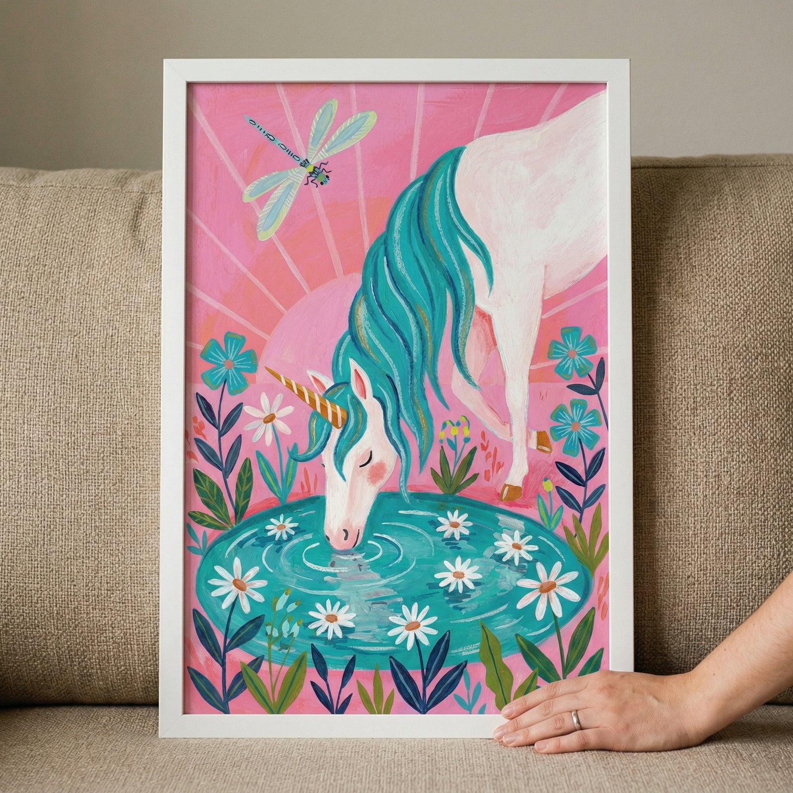 A framed illustration shows a white mythical creature with a single golden horn drinking from a turquoise pond. It has a long, flowing teal mane. Small white flowers with yellow centers float in the water, and various blue and white flowers grow around the edge. The background is a vibrant pink sky with a sunburst effect. A dragonfly with teal and green wings flies in the upper portion. The artwork is held in a white frame on a neutral couch, with a person's hand resting on the corner.