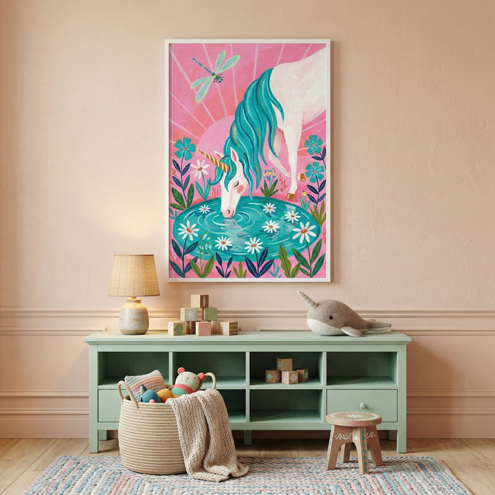 Colorful unicorn illustration with a teal mane drinking from a pond surrounded by flowers on a bright pink background. The framed artwork hangs above a mint green shelf in a playful nursery setting with toys, adding a whimsical fantasy touch to the space.
