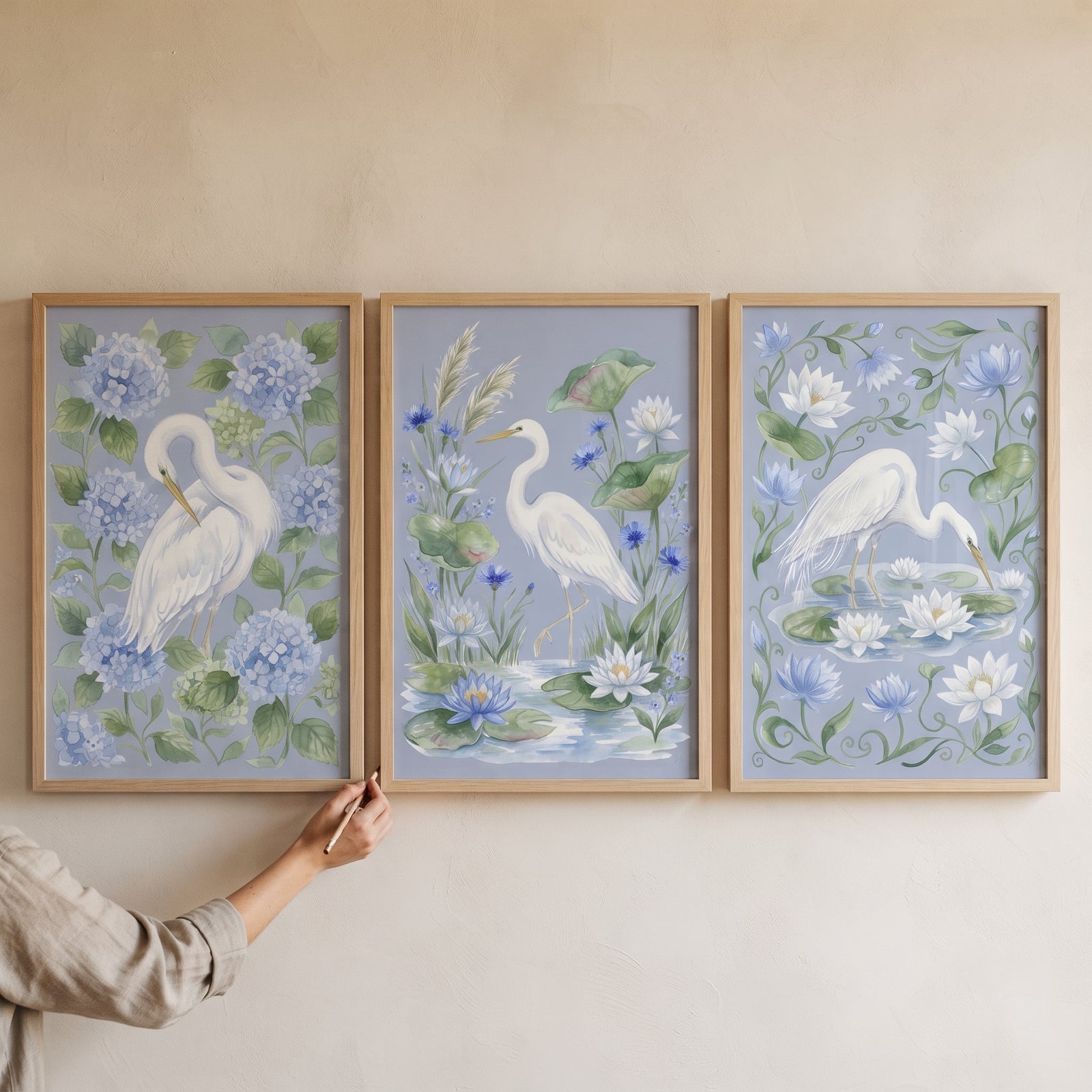 A hand adjusts a powder blue botanical heron bird wall art set of 3 featuring white herons and soft blue flowers.