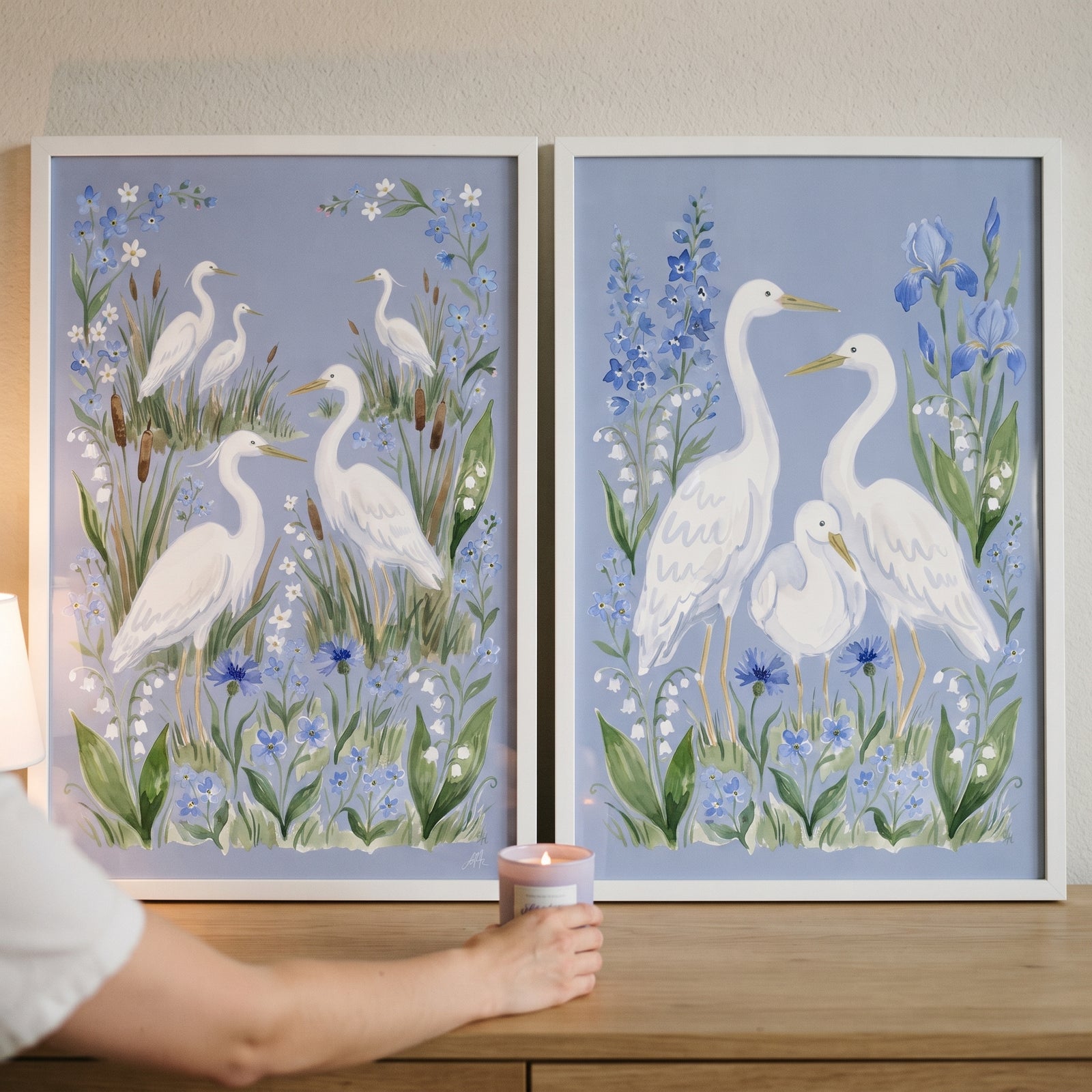 A hand reaches for a candle in front of a powder blue wildflower egret bird wall art set of 2 on a wooden surface.
