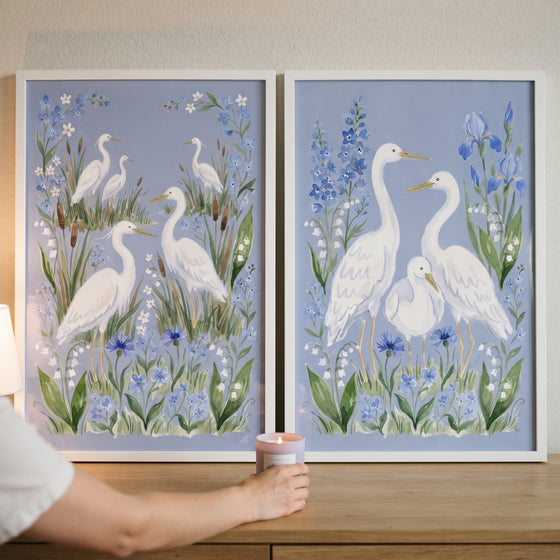 A hand reaches for a candle in front of a powder blue wildflower egret bird wall art set of 2 on a wooden surface.