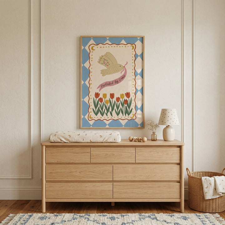 Minimalist nursery with a wooden dresser and soft neutral decor beneath a framed print of a bird carrying a ribbon that reads "Protected By Grace" above a row of tulips. The artwork adds a gentle and comforting focal point to the calm, light filled space.