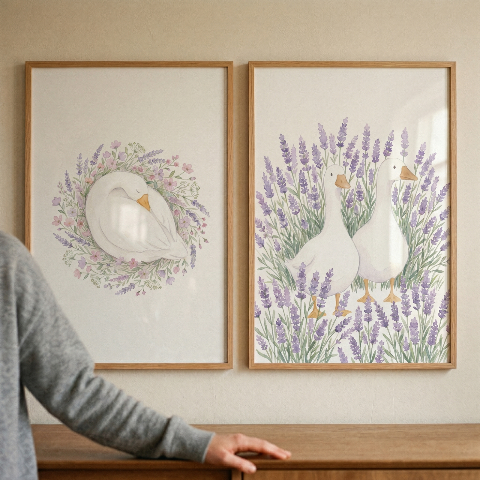 Two framed watercolor illustrations show white ducks in soft floral scenes one resting curled inside a wreath of pastel flowers and the other standing among tall lavender stems. A person’s arm rests on the wooden surface below adding a warm lived in touch to the calm nature inspired nursery display.